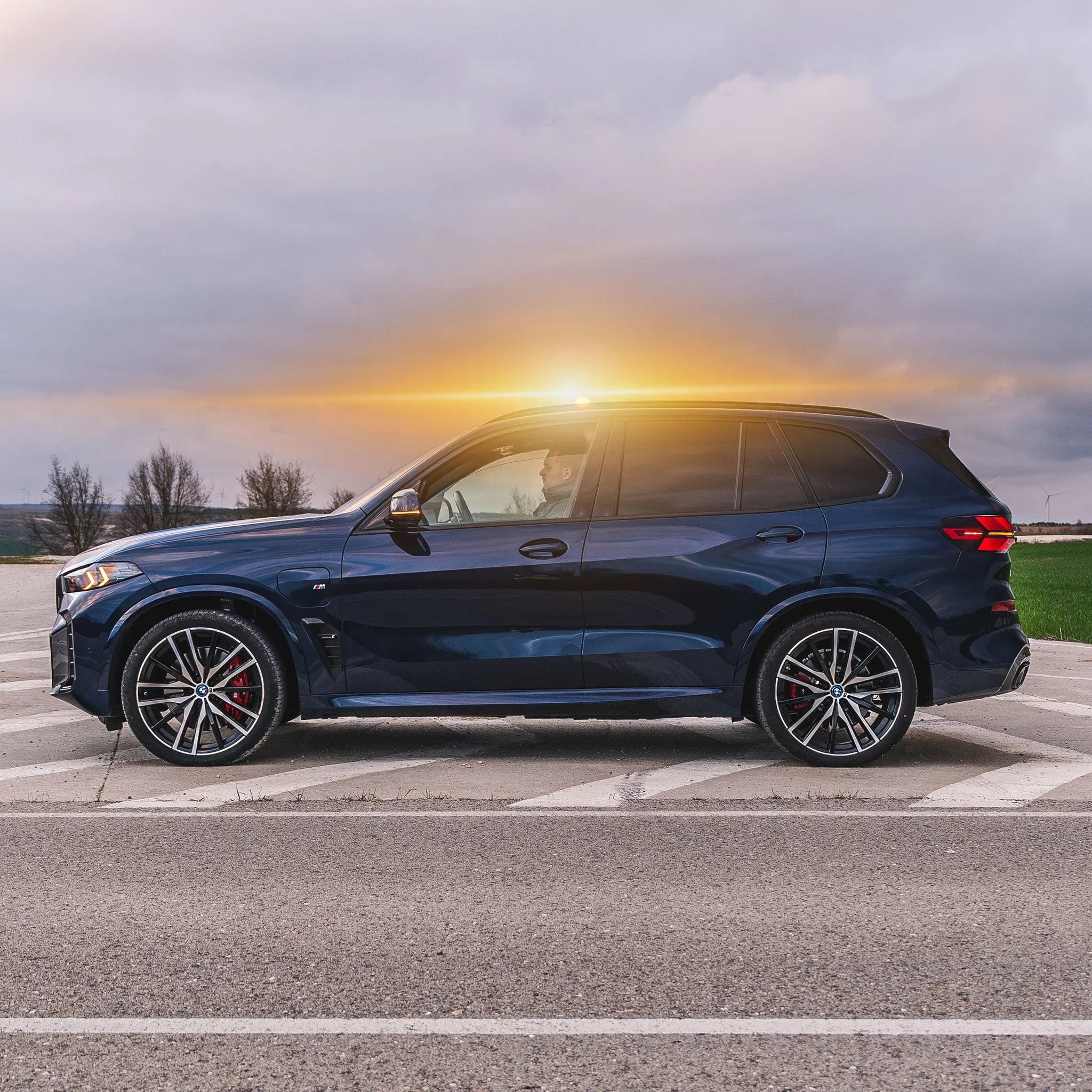 SUV BMW M oscuro con conductor visible al atardecer en carretera asfaltada con paisaje rural