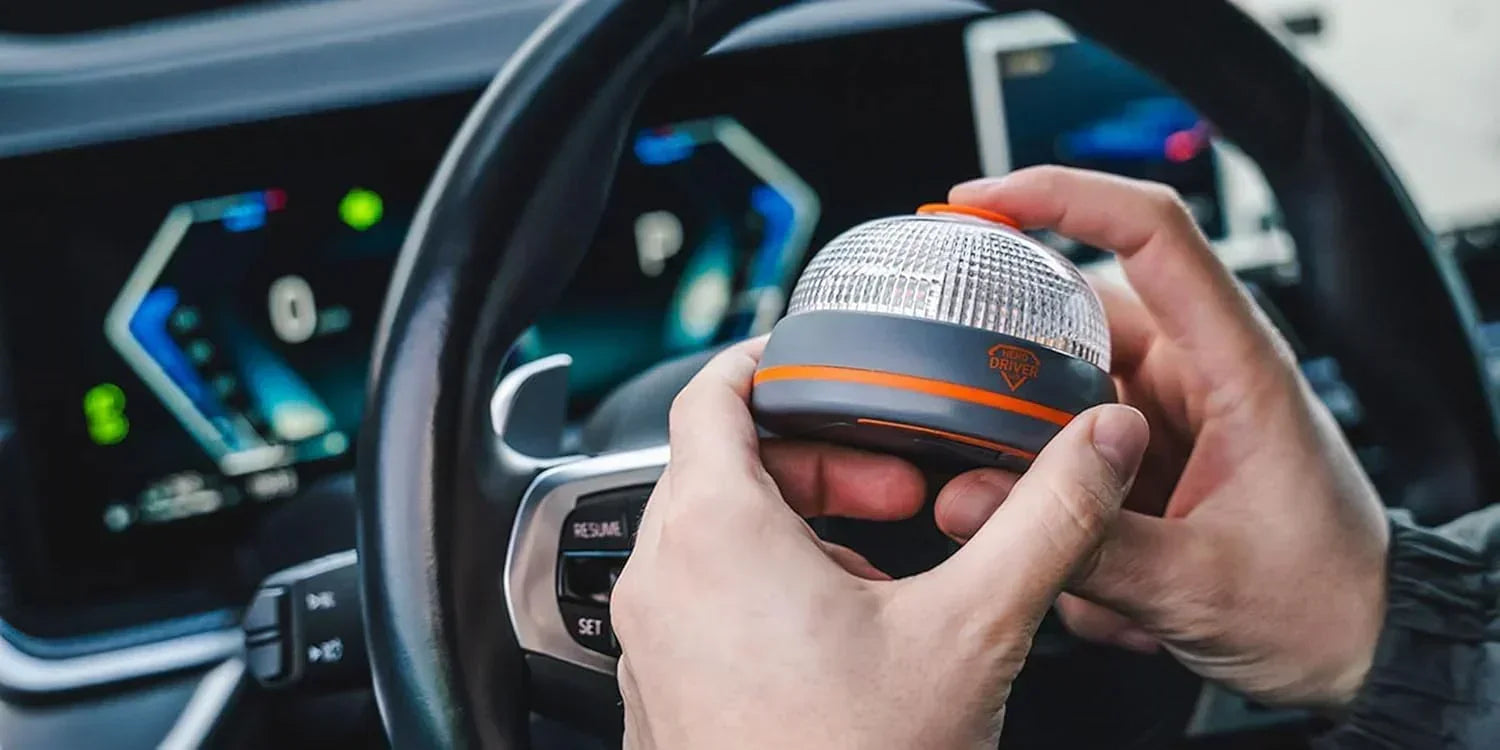 Manos sosteniendo baliza de emergencia LED con botón naranja dentro de un auto moderno frente al volante