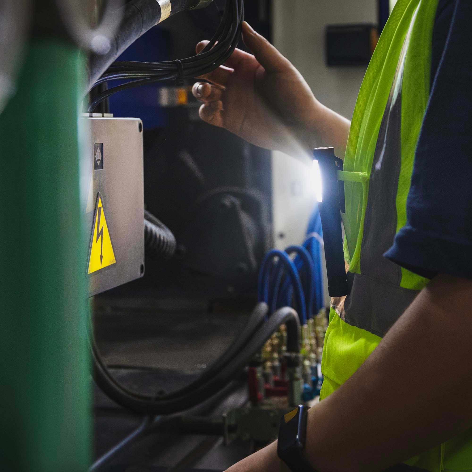 Trabajador con chaleco reflectante usando linterna LED en máquina industrial con cables y señal de advertencia eléctrica