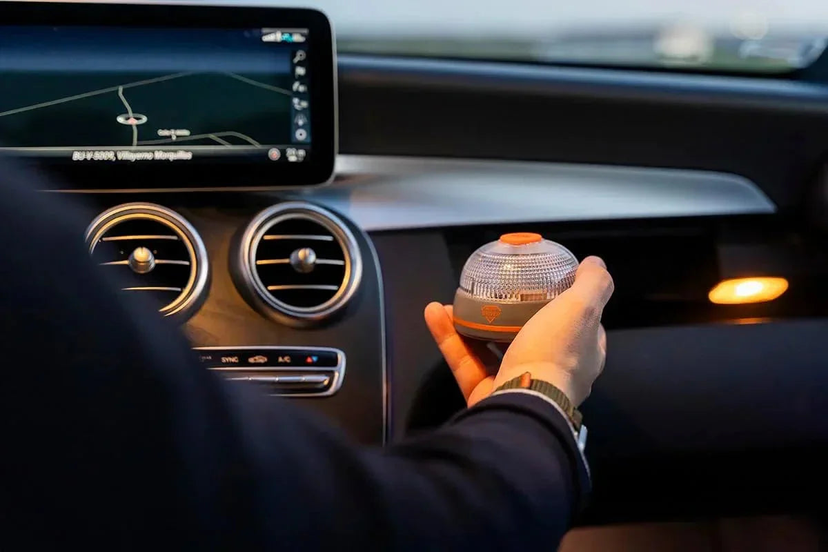 Interior de coche con mano sosteniendo baliza LED homologada DGT junto a pantalla de GPS y aire acondicionado