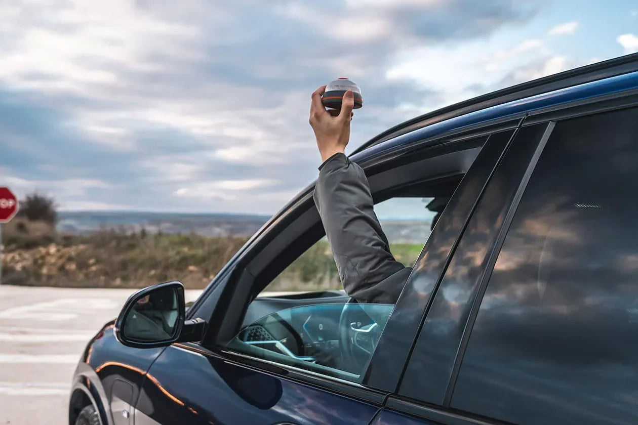 Brazo extendido desde ventana de coche azul sosteniendo una baliza geolocalizada en campo abierto
