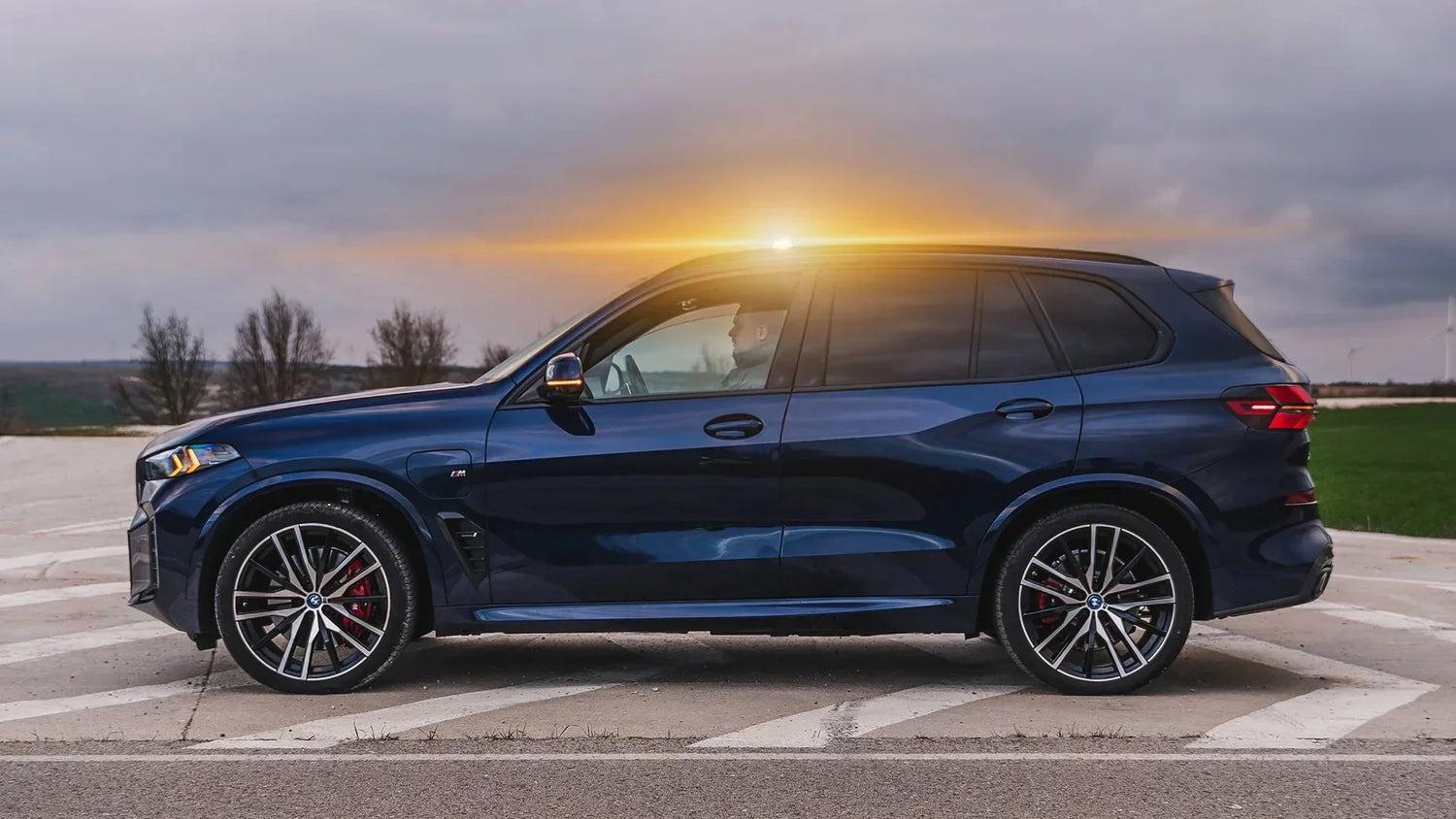 SUV BMW X5 M azul oscuro estacionado en carretera con conductor y luces delanteras encendidas