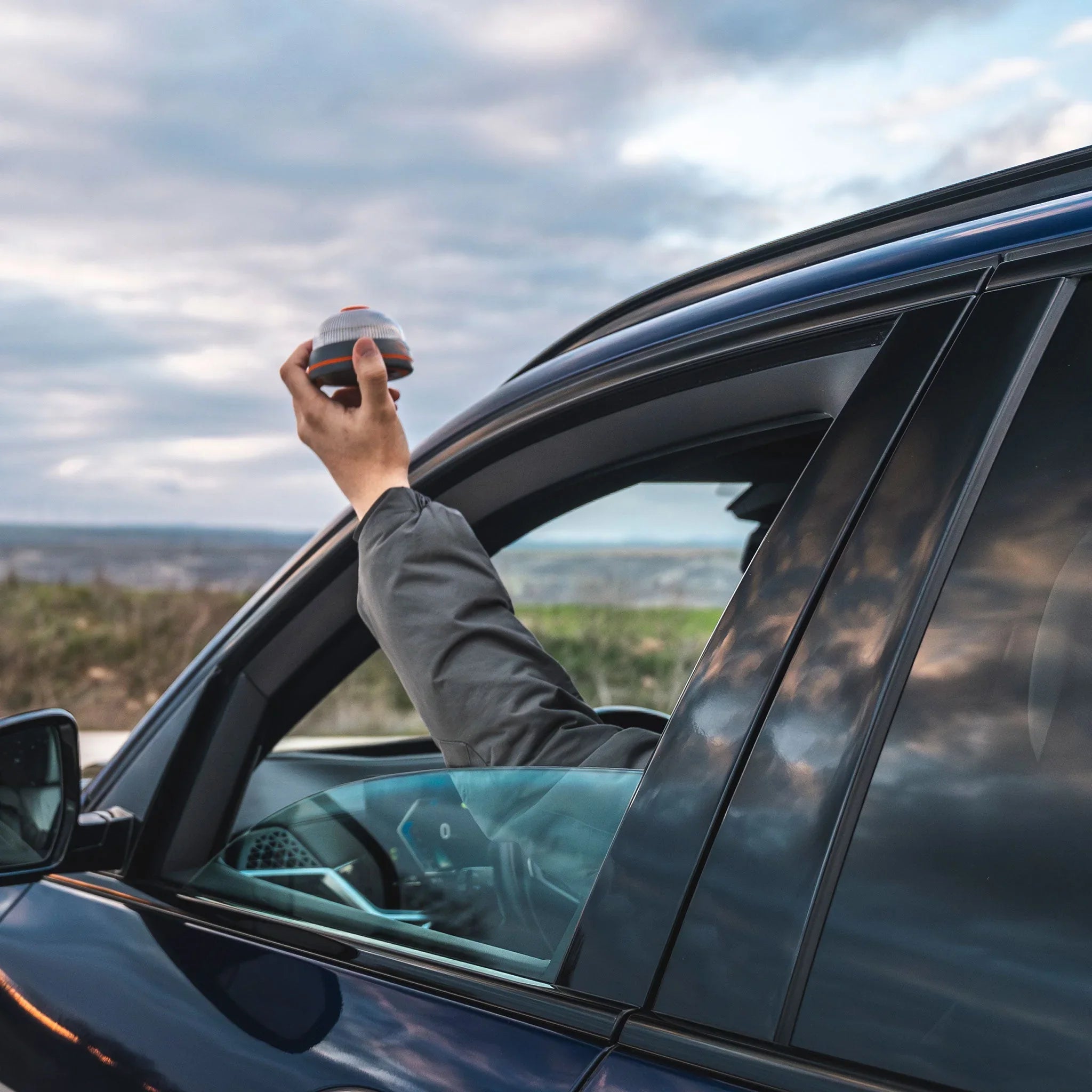 Brazo extendido desde ventana de coche sosteniendo dispositivo electrónico pequeño en exterior nublado